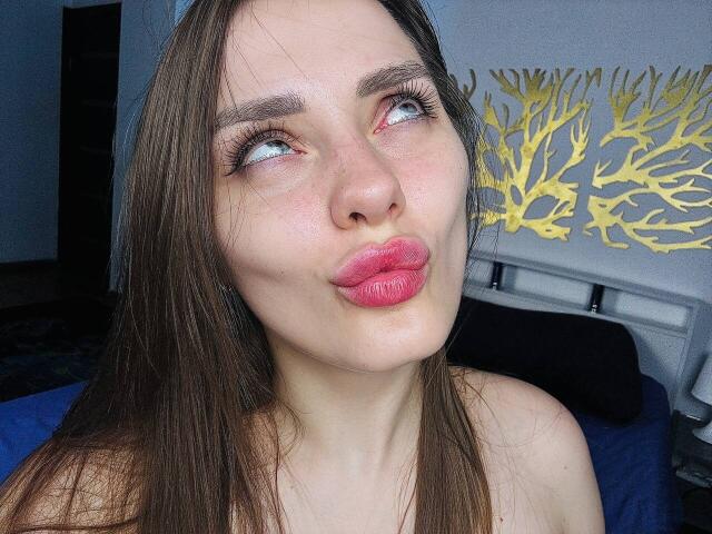 TenderAmberr, met volle, roze lippen, bruin, steil haar en blauwe ogen, kijkt omhoog. Ze heeft dikke, zwarte wimpers en een lichte huidskleur. Haar tepels zijn niet zichtbaar..