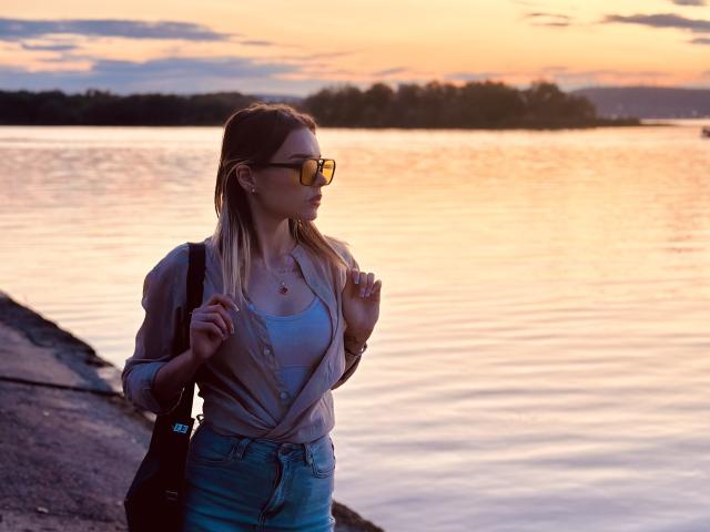 EmelyHailey with blonde hair, wearing yellow sunglasses, a pink shirt, and blue jeans, standing by a waterway at sunset, holding a black bag, hands near chest.