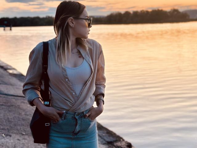 EmelyHailey with tits visible in a beige button-up shirt, white tank top, blue jeans, black bag, and sunglasses, standing by a lake at sunset. Hands in pockets.