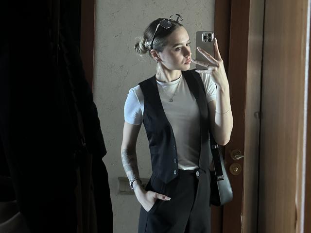 EmelyHailey with hands in black vest pockets, white t-shirt, black pants, and hands in pockets, taking mirror selfie, wearing glasses on head, in dimly lit room.