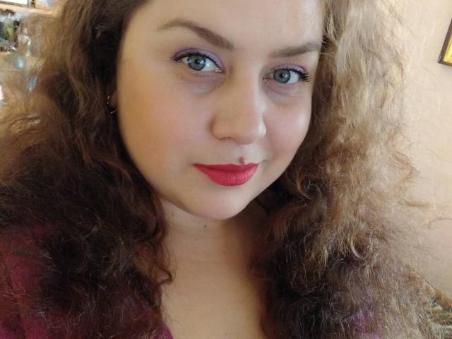 SunshineMiranda with curly brown hair, blue eyes, purple eyeliner, red lipstick, and a slight smile. She's wearing a red top, looking cute and attractive.
