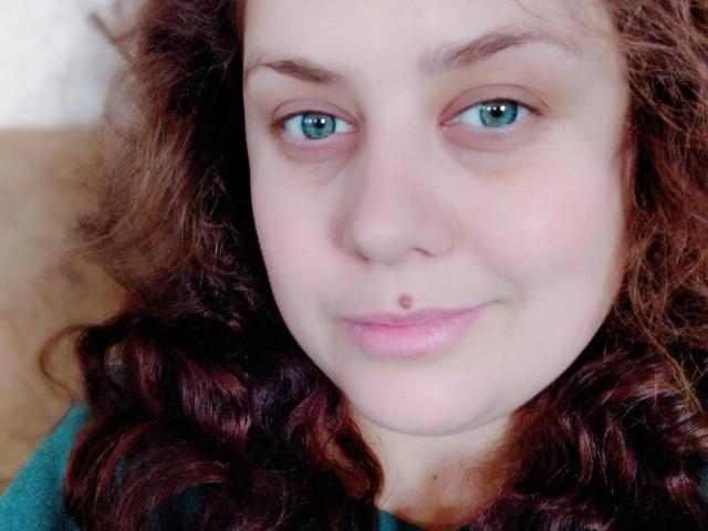 SunshineMiranda with curly brown hair, blue eyes, and a small beauty mark below her upper lip, smiling softly. She's wearing a teal shirt, looking relaxed.