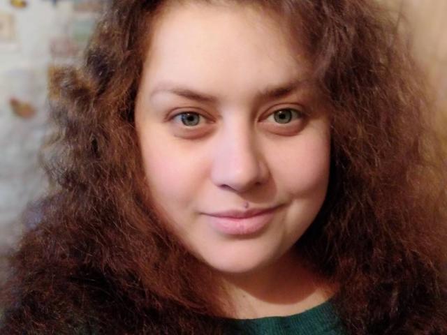 SunshineMiranda with curly brown hair, green eyes, and a subtle smile, wearing a green top, has a small nose ring in her right nostril.