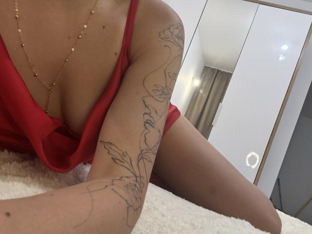 EricaLust in a red dress, showing a tattooed arm with floral designs, gold necklace, and her tits slightly visible, lying on a white fluffy surface.