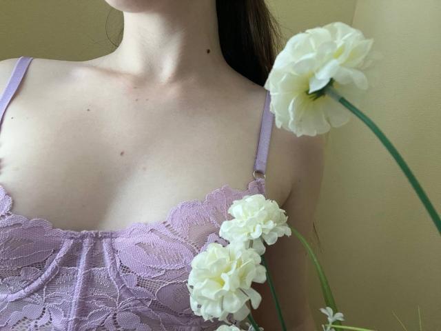 Valissia's pale boobs in a lilac lace bra, with white flowers in the foreground, her neck and a small mole visible, brown hair partially in frame.