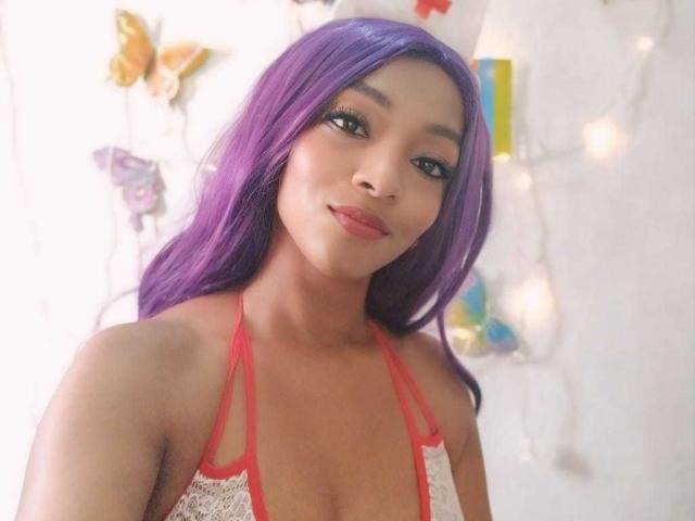 BrendaReyna with purple hair, red halter top, showing her tits, smiling at the camera, looking cute with eyeliner and lipstick. Soft light, slight blur in the background.