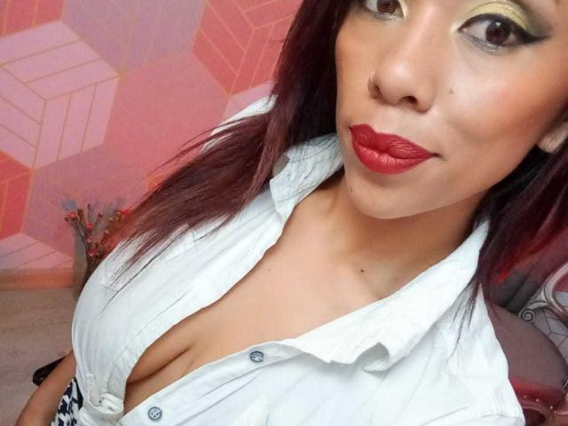 AmmyTorrezX with red lipstick, eye makeup, and white shirt, showing cleavage, has medium tits, brown skin, and red-brown hair, in a selfie, smiling slightly.