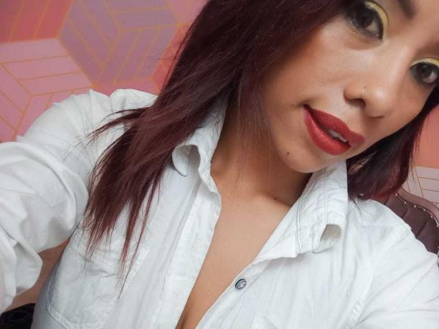 AmmyTorrezX, com batom vermelho, piercing no nariz e camisa branca de botões, mostrando um pouco o colo, tem cabelos longos e escuros e uma sardinha fofa no queixo..