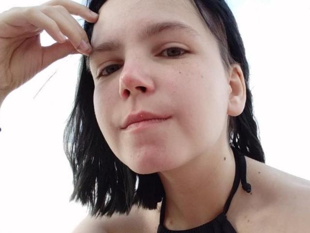 SpicyDora with black shoulder-length hair, slightly parted lips, fair skin, and a subtle smile, touching her forehead with a hand, wearing a black halter top.