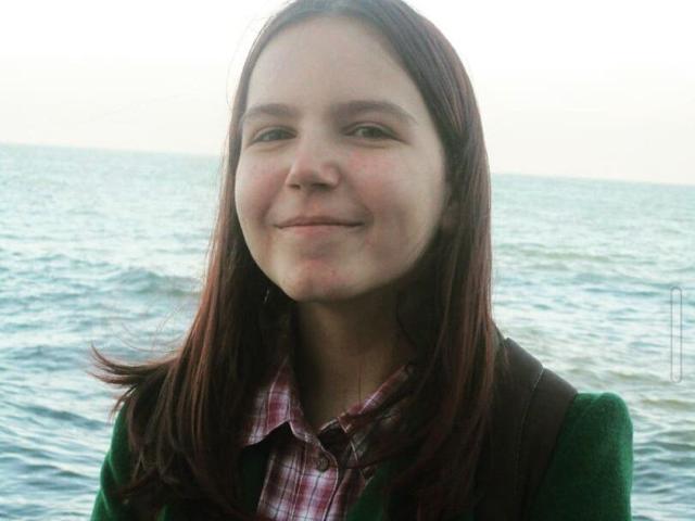 SpicyDora has a cute smile, light skin, and medium brown hair, wearing a green sweater over a pink plaid shirt, standing by the blue ocean.