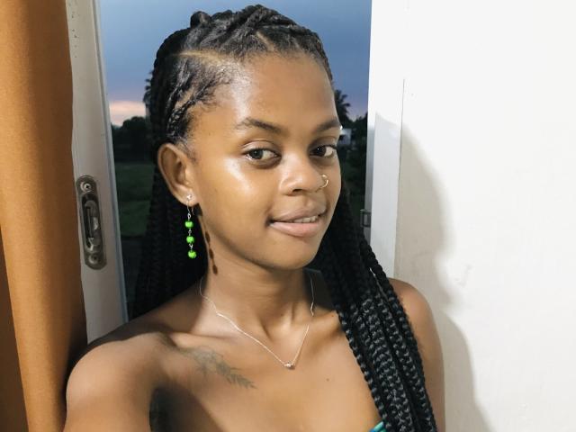 JessiccaHot has dark brown skin, long black braids, green bead earrings, nose ring, small tattoo on left shoulder, and a subtle smile, standing by a window at dusk.