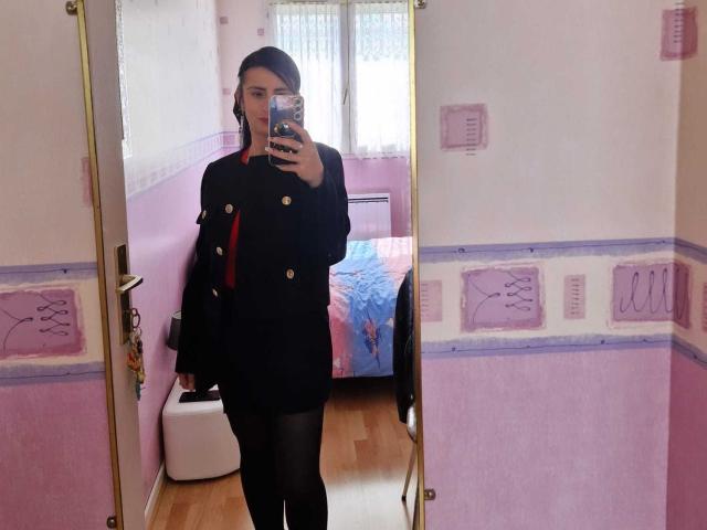 BrendaCantorel stands in front of a mirror wearing a black double-breasted coat, black tights, and a red scarf, holding a phone to take a mirror selfie.