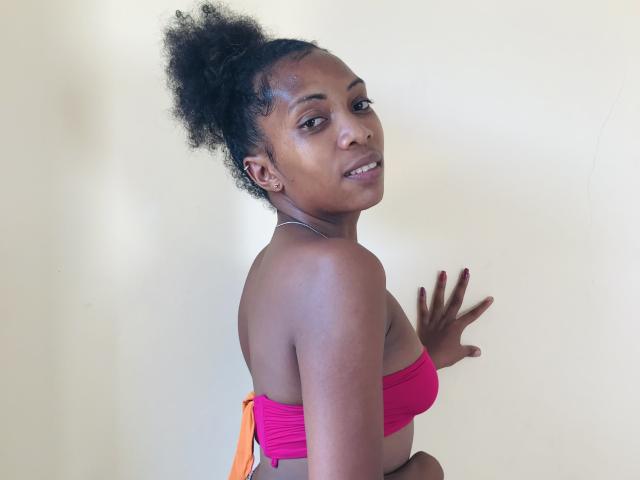 AntoniaMagik with dark skin, curly black hair in a bun, wearing a pink strapless top, showing her small tits, looking over her shoulder, hand on a white wall.