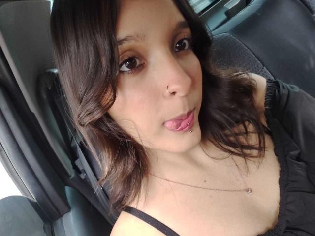 LanaKlaire with dark wavy hair, brown eyes, nose ring, and pink tongue piercing, wearing a black top, sitting in a car. Her tits are small and she's looking at the camera.