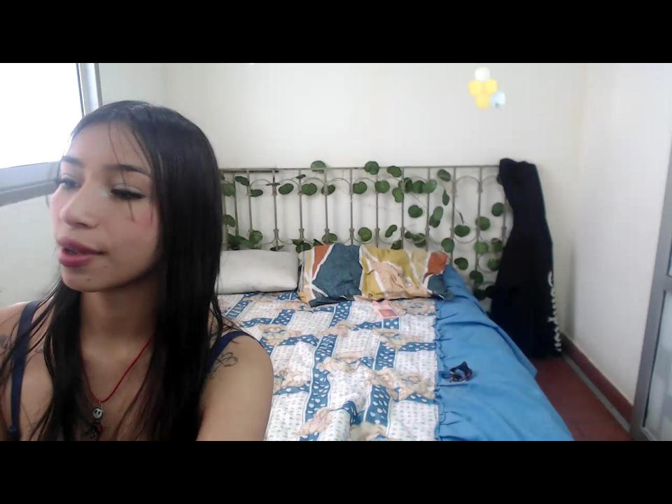 PervertHotSex with long black hair, wearing a blue tank top, standing by a bed with a patterned quilt, a pillow, and green leaves on the headboard. Her ass is unseen.