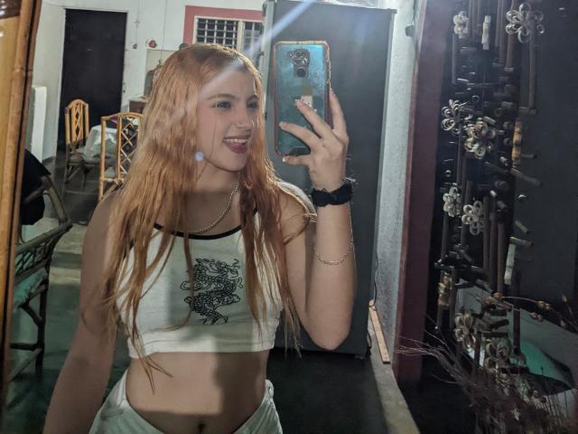 JeyLove with wet red hair, smiling, holding a phone, wearing a white crop top with black print, showing her tits, in a mirror selfie, in a dim room.