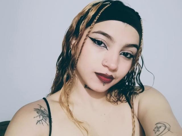 JeyLove with wet blonde braids, dark lipstick, eyeliner, nose ring, and lip piercing, showing tattoos on shoulders, wearing black headband and top, looking at camera.