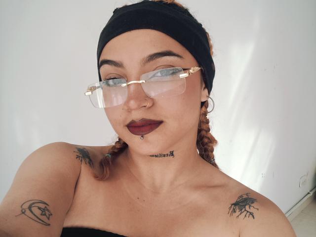 JeyLove with glasses, black headband, and dark lipstick, showing tattoos on shoulders and neck, braided hair, nose ring, and a chin piercing, smiling at the camera.