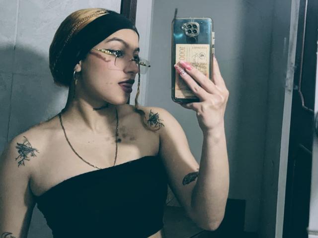 JeyLove with glasses, black tube top, tattoos on shoulders, pink nails, headwrap, holding phone, looking in mirror, medium skin, small breasts, earbuds, dark lipstick, subtle makeup.