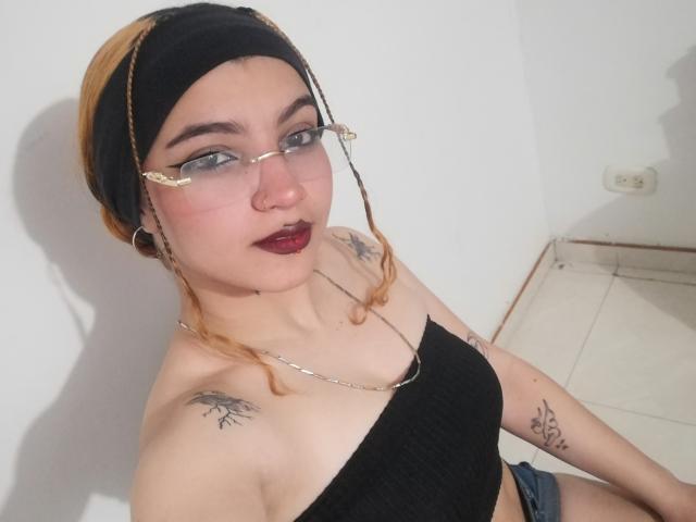 JeyLove with glasses, black headband, and red lipstick, showing off her tits in a black top, tattoos on shoulders, and blue shorts, sitting on a tiled floor.