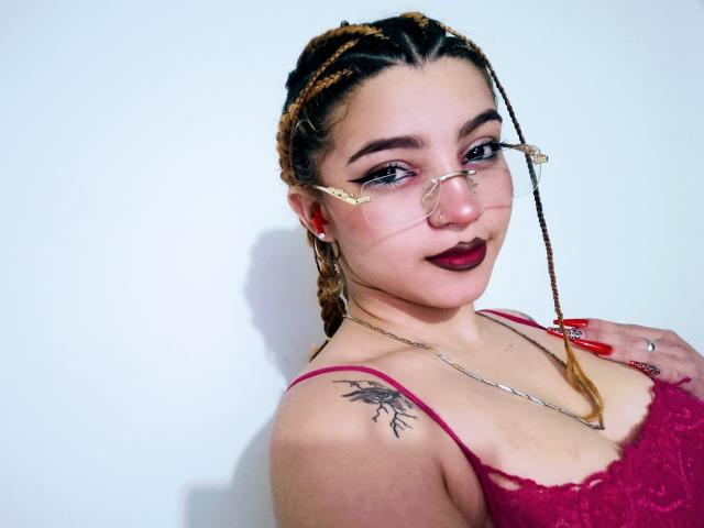 JeyLove with braided hair, glasses, red lipstick, and a tattoo on her shoulder, wearing a red lace top, showing off her tits and red nails.
