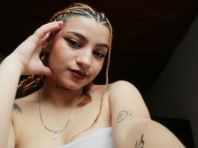 JeyLove with braided orange hair, dark lipstick, and white strapless top, showing tattoos on neck and arm, hand on head, looking at camera with slight smile.