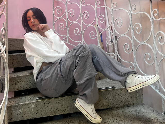 AitanaJohns está sentada en unos escalones de hormigón, vestida con una camisa blanca, pantalones grises holgados y zapatillas blancas, con el mentón apoyado en la mano, mirando hacia abajo..