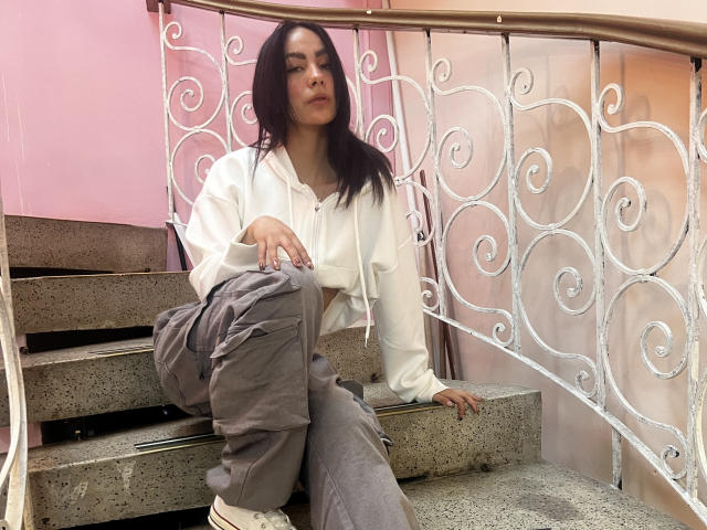 AitanaJohns, mit langen, dunklen Haaren, sitzt auf einer Betontreppe, trägt eine weiße Kapuzenjacke, graue Hosen und weiße Turnschuhe und blickt mit einem leichten Lächeln in die Kamera..