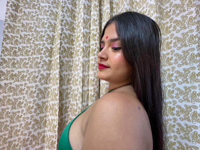 NayaYalitza with long black hair, red lipstick, green top, nose piercing, red dot on forehead, side profile, looking down, patterned curtain, light skin, slight smile, small tattoo on shoulder.