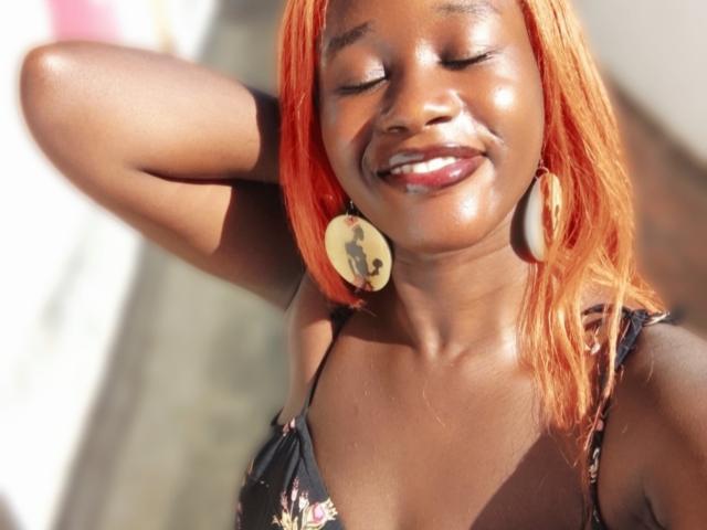 BustyBae with shiny brown skin and bright orange hair, smiling with eyes closed, wearing a floral dress and large wooden earrings, sunlight highlighting her glossy face.