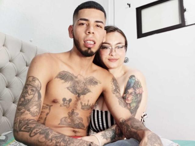 MiaAndLeon, with tattoos, sits on bed, arms around chest of shirtless Leon with eagle tattoo, Leon's hand on Mia's ass, Mia's tits visible, both smiling.