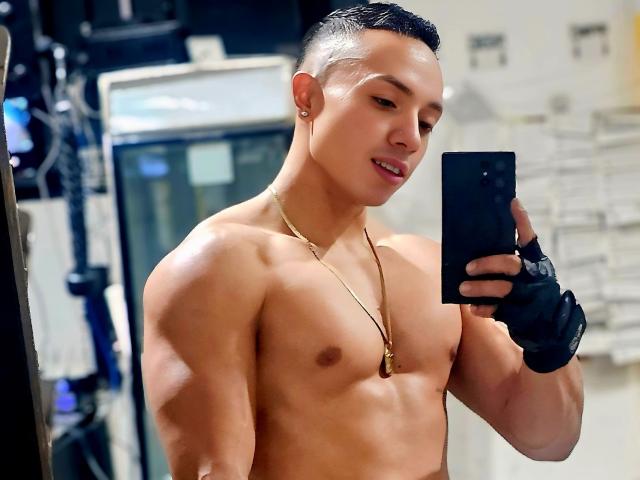 Shirtless DamianLove69 with a toned, muscular build, short black hair, gold necklace, black glove, holding a phone, taking a mirror selfie, smiling, showing defined abs and chest.