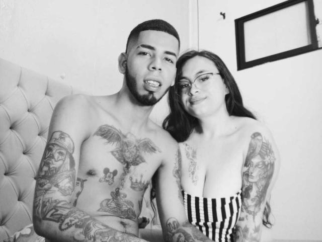 Black-and-white photo of MiaAndLeon with tattoos, topless Leon has eagle chest tattoo, bearded, MiaAndLeon with glasses, striped dress, large tits, arm tattoos, smiling, Leon's arm around MiaAndLeon.