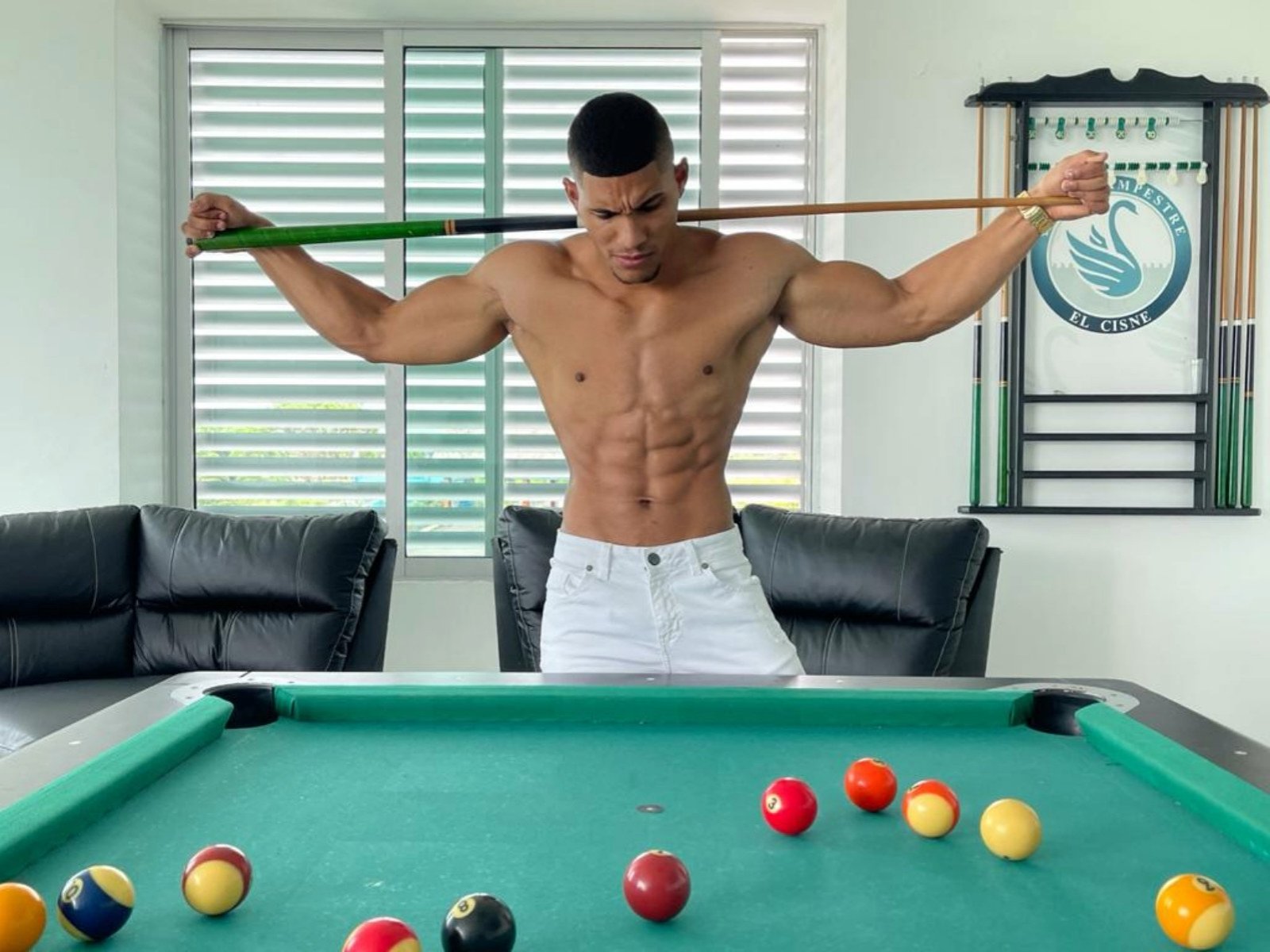 TeddyFit, sem camisa, musculoso, com pele bronzeada, em pé sobre uma mesa de bilhar verde, segurando um taco de bilhar, vestindo calças brancas, com várias bolas de bilhar coloridas na frente..