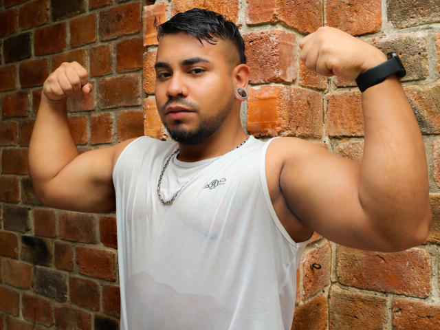 BrianMarly, muscular, flexing biceps, wearing a white, semi-transparent tank top, showing nipples, black wristband, ear gauges, short dark hair, and a beard, standing against a brick wall.