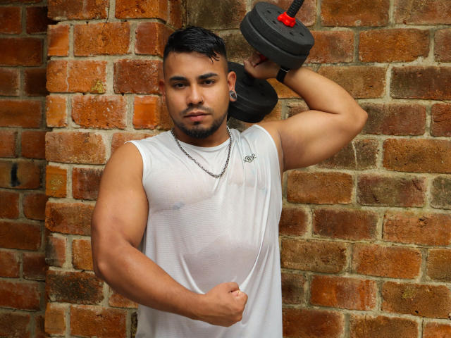 BrianMarly with a muscular build, short black hair, and a white sleeveless shirt, flexing his arm while holding a dumbbell against a brick wall. Visible nipple.
