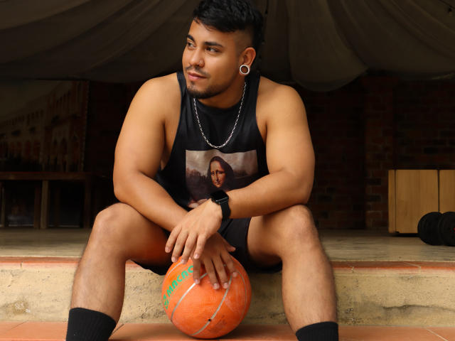 BrianMarly, muscular, tan-skinned, black hair, small ear piercings, black tank top with photo, black shorts, sitting on brick steps, orange basketball, black socks, silver chain, black wristband.
