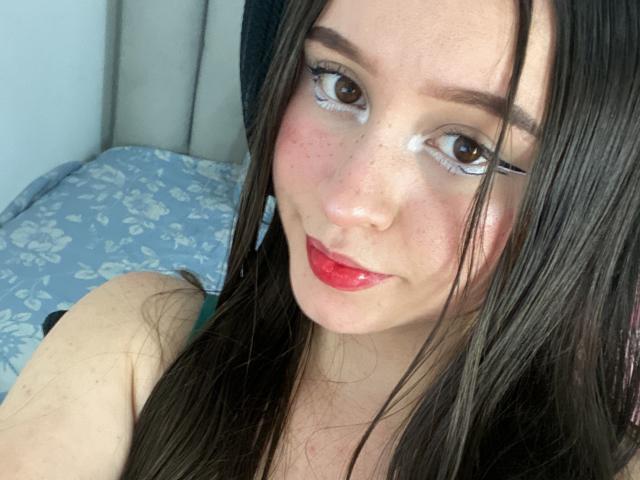 SophieHale with long brown hair, red lipstick, and white eye makeup, smiling at the camera, showing freckles on her face, with her tits barely visible in the frame.