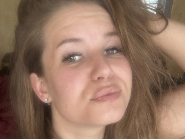 BertieMyra's face, with light brown hair, blue eyes, and subtle pout, wearing small diamond earrings, has slightly flushed cheeks and a relaxed, confident expression.