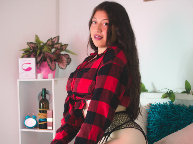 Amy Phillips, com cabelos castanhos longos, camisa xadrez vermelha e preta amarrada na frente, calcinha de rede preta, sorrindo para a câmera, sentada em uma cama com um travesseiro azul..