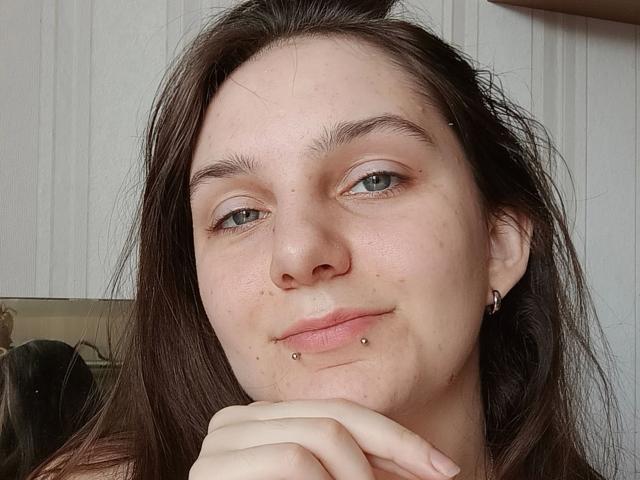 MayaSchatz-hot with brown hair, green eyes, nose piercing, lip piercing, and earrings, smiling slightly with hands under chin. Her skin is fair with some acne.