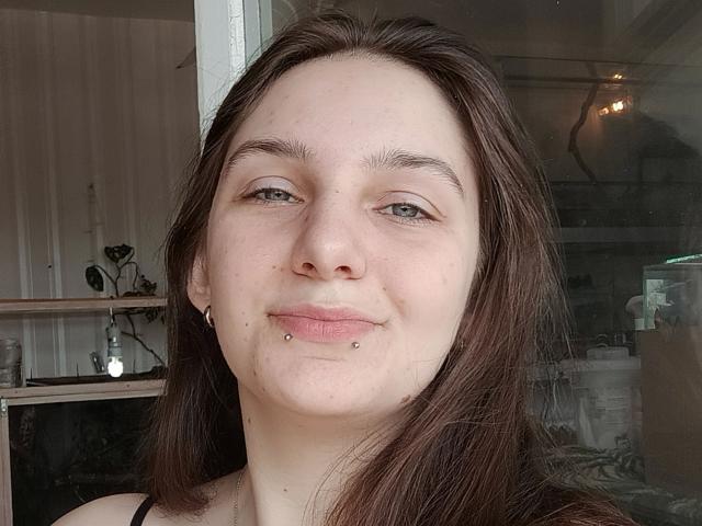 Maya Schatz, com cabelo castanho, pele clara, olhos verdes e um piercing labret no lábio inferior, sorrindo levemente. Ela está usando pequenos brincos de prata e uma blusa preta..