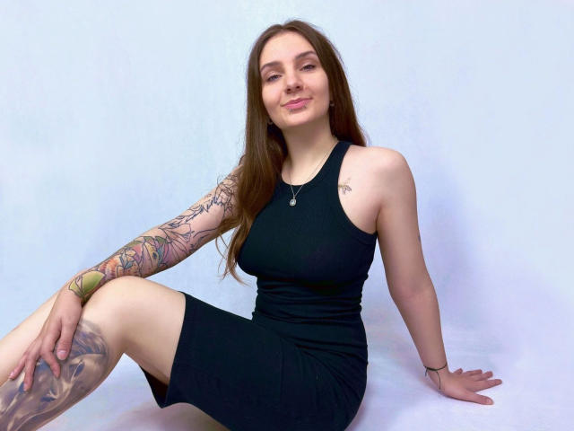 MayaSchatz – attraktiv mit langen braunen Haaren, schwarzem trägerlosen Kleid, Tattoos am Arm und Oberschenkel, sitzend, lächelnd, Hände auf dem weißen Boden, weißer Hintergrund, schlank, kleine Brüste, süß, lässig..