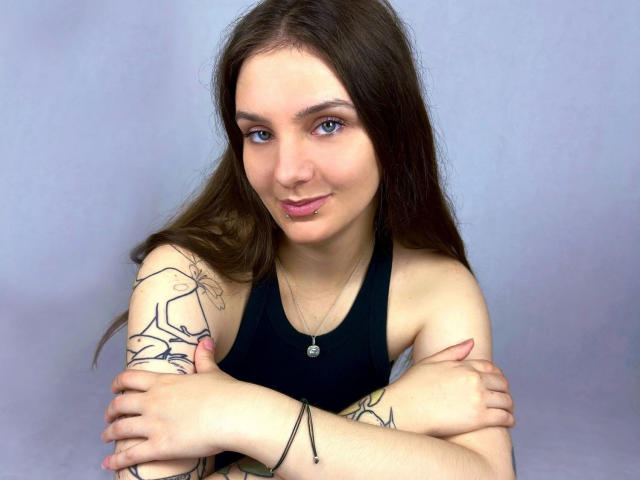 Maya Schatz, atraente, com longos cabelos castanhos, olhos azuis e um piercing no lábio, cruza os braços sobre tatuagens, vestindo uma blusa preta de alças e um colar de prata, sorrindo levemente..
