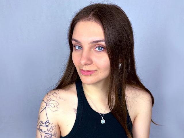 MayaSchatz-hot tem cabelos castanhos longos, olhos azuis, um piercing rosa no lábio, uma camiseta preta, uma tatuagem floral no ombro direito e um colar de prata, tudo isso em um fundo cinza simples..