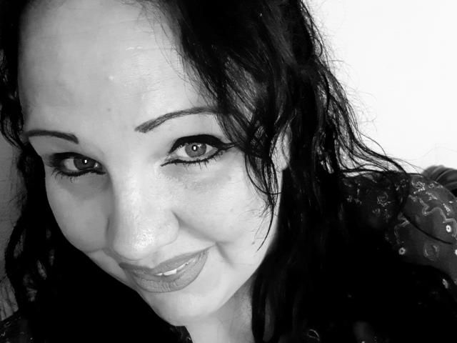 Blackbeauty43-hot has curly black hair, light skin, and is smiling with dark eyeliner and glossy lipstick. She's wearing a patterned shirt, and her eyes are looking up.