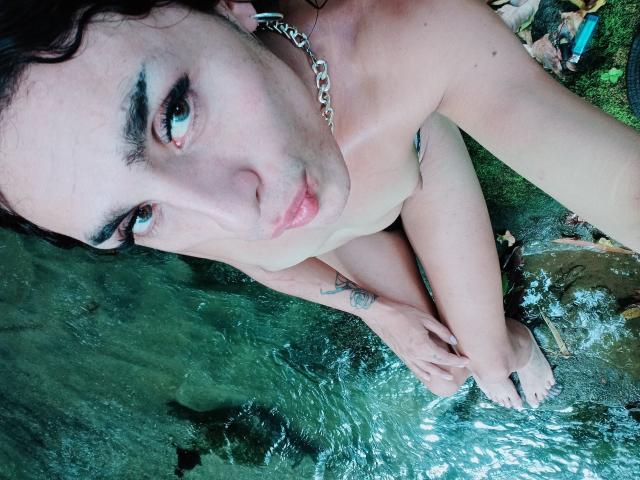 Crystalinn, naked, with small tits, crouching in clear water, looking up at the camera, dark curly hair, chain necklace, tattoo on left arm, bare feet, green water.