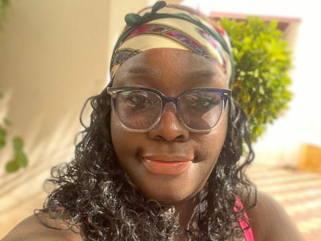 TooMuchSquirts a la peau foncée, des cheveux noirs bouclés, porte un foulard coloré et de grandes lunettes bleues. Elle sourit, son visage expressif baigné de soleil..
