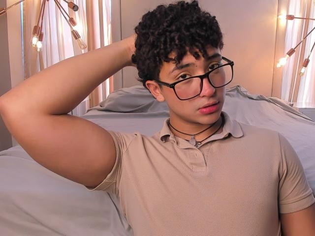 Valentin69s with curly black hair, glasses, pink lipstick, beige polo, and necklace, raising arm, looking at camera, lying on bed with white sheets, string lights in background.