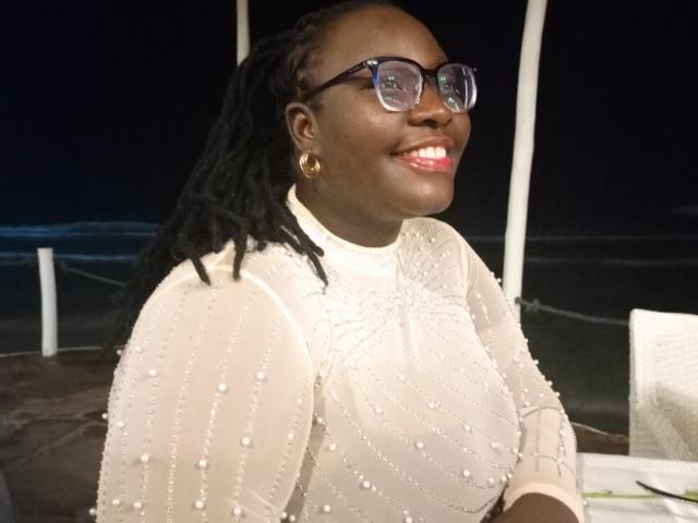TooMuchSquirts, dark-skinned, curvy, wearing glasses, sheer white dress with bead patterns, gold hoop earrings, black dreadlocks, smiling with red lipstick, nighttime, visible breast outline.